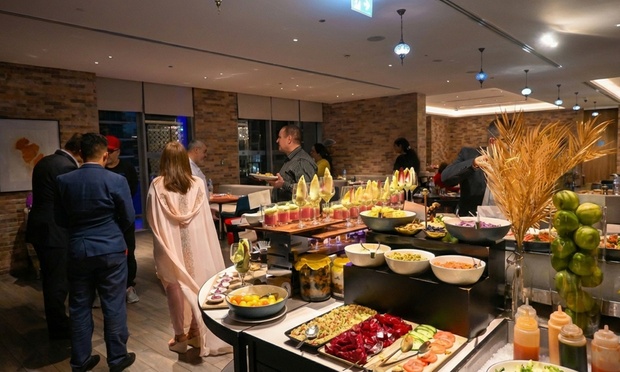 A beautifully arranged buffet spread with a variety of international dishes and a traditional OUD instrument in the background, set in a restaurant dining area with both indoor and outdoor seating visible.